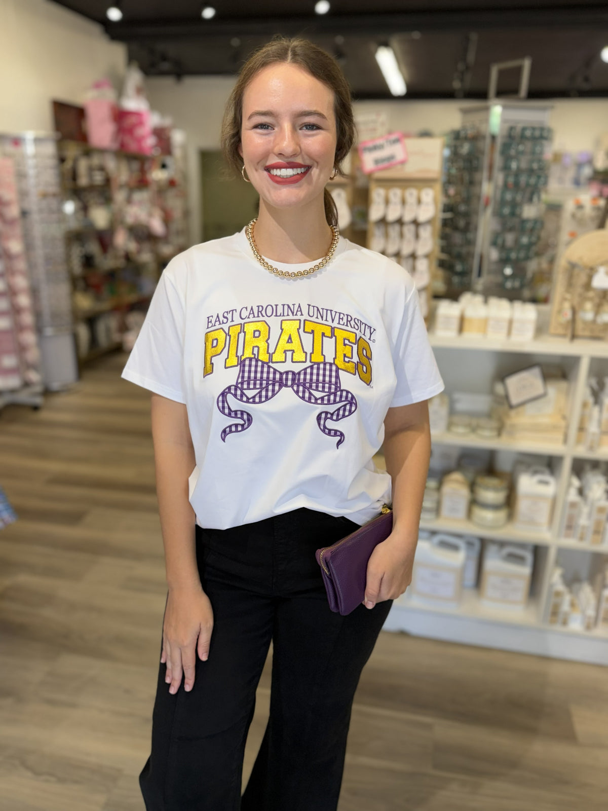 This is a white short-sleeve graphic T-shirt featuring an East Carolina University Pirates design. The front reads “EAST CAROLINA UNIVERSITY” in purple above oversized “PIRATES” lettering in gold with purple outlines. A large purple gingham bow graphic with flowing ribbon tails adds a cute, feminine touch beneath the collegiate lettering. The relaxed crewneck silhouette gives it a casual, spirited look that’s perfect for game days, campus wear, or everyday outfits.