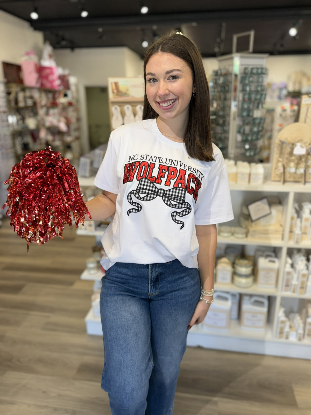 This is a white short-sleeve NC State University graphic T-shirt with a classic crew neckline and relaxed fit. The front features “NC STATE UNIVERSITY” in black above oversized “WOLFPACK” lettering in bold red with black detailing. A large black-and-white gingham bow with flowing ribbon tails adds a cute, feminine touch to the collegiate design. It’s a fun game-day tee that pairs classic school spirit with a trendy bow detail.