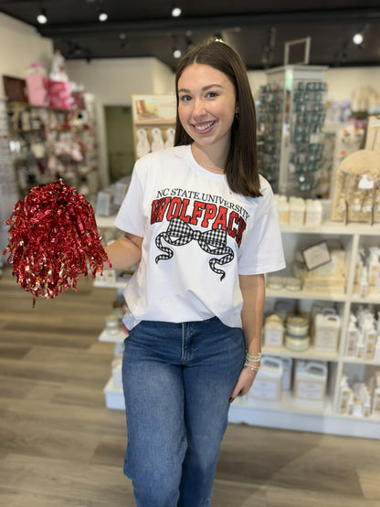 This is a white short-sleeve NC State University graphic T-shirt with a classic crew neckline and relaxed fit. The front features “NC STATE UNIVERSITY” in black above oversized “WOLFPACK” lettering in bold red with black detailing. A large black-and-white gingham bow with flowing ribbon tails adds a cute, feminine touch to the collegiate design. It’s a fun game-day tee that pairs classic school spirit with a trendy bow detail.