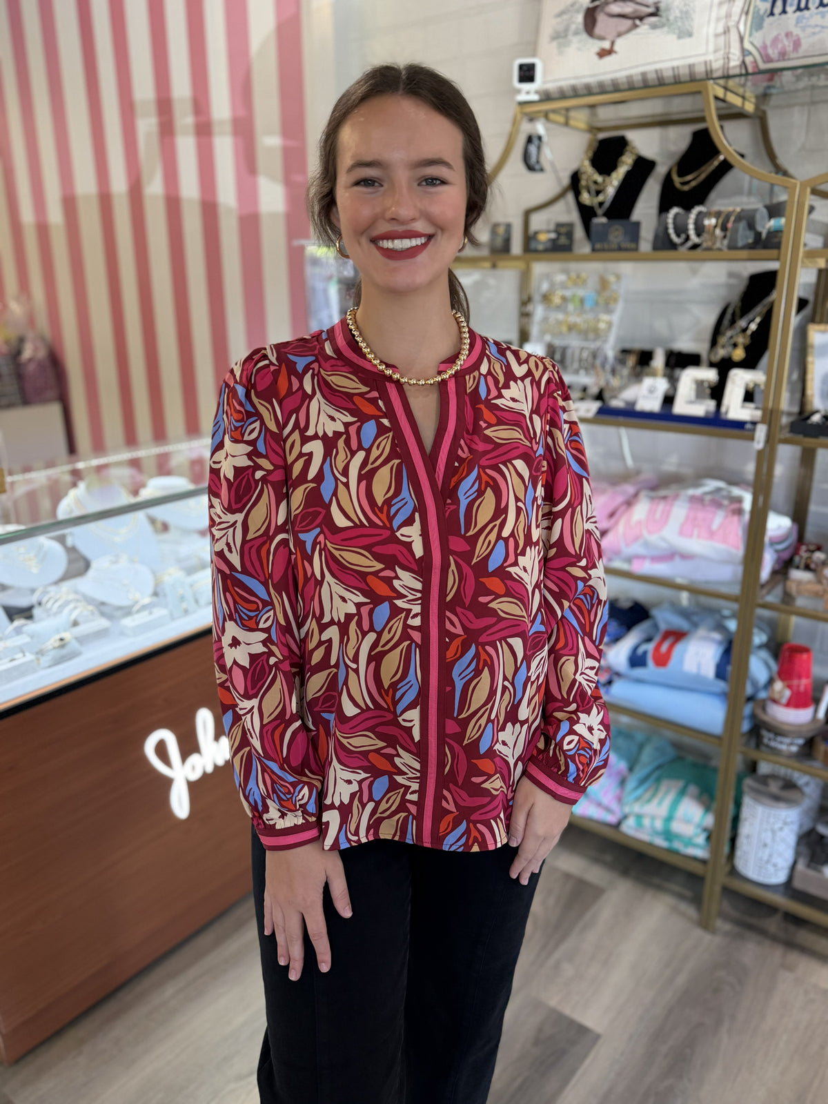 This is a long-sleeve printed blouse featuring a bold abstract botanical pattern in rich burgundy, raspberry pink, cream, rust, tan, and pops of light blue. It has a split V-neckline accented with contrasting pink trim that continues down the front for a polished finish. The relaxed silhouette is complemented by slightly gathered shoulders and softly puffed sleeves, finished with coordinating trimmed cuffs. The vibrant print and flowy fit make it an eye-catching piece that pairs beautifully with black pants