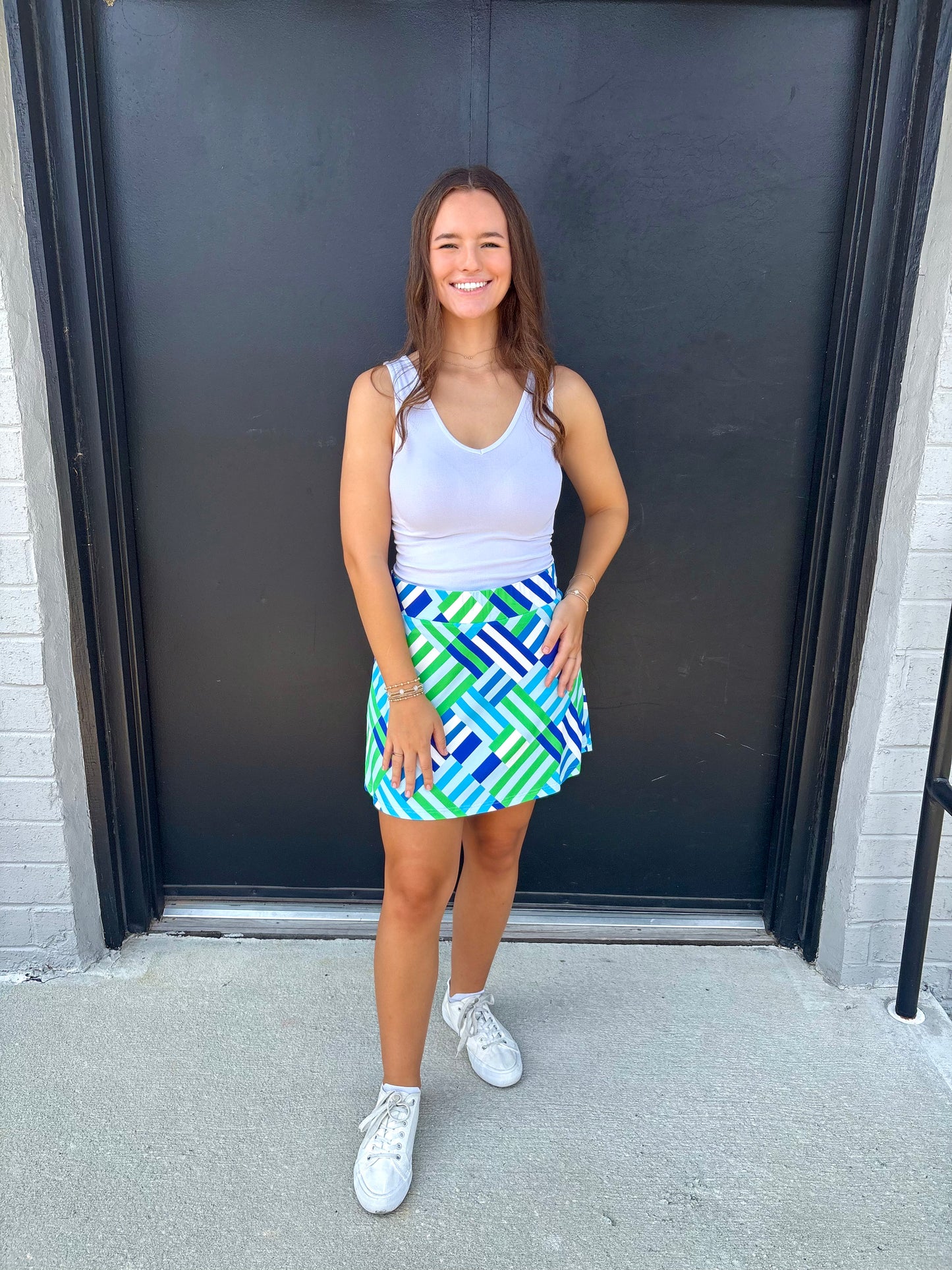 Woman wearing a white tank top and colorful geometric skirt standing in front of a black door.