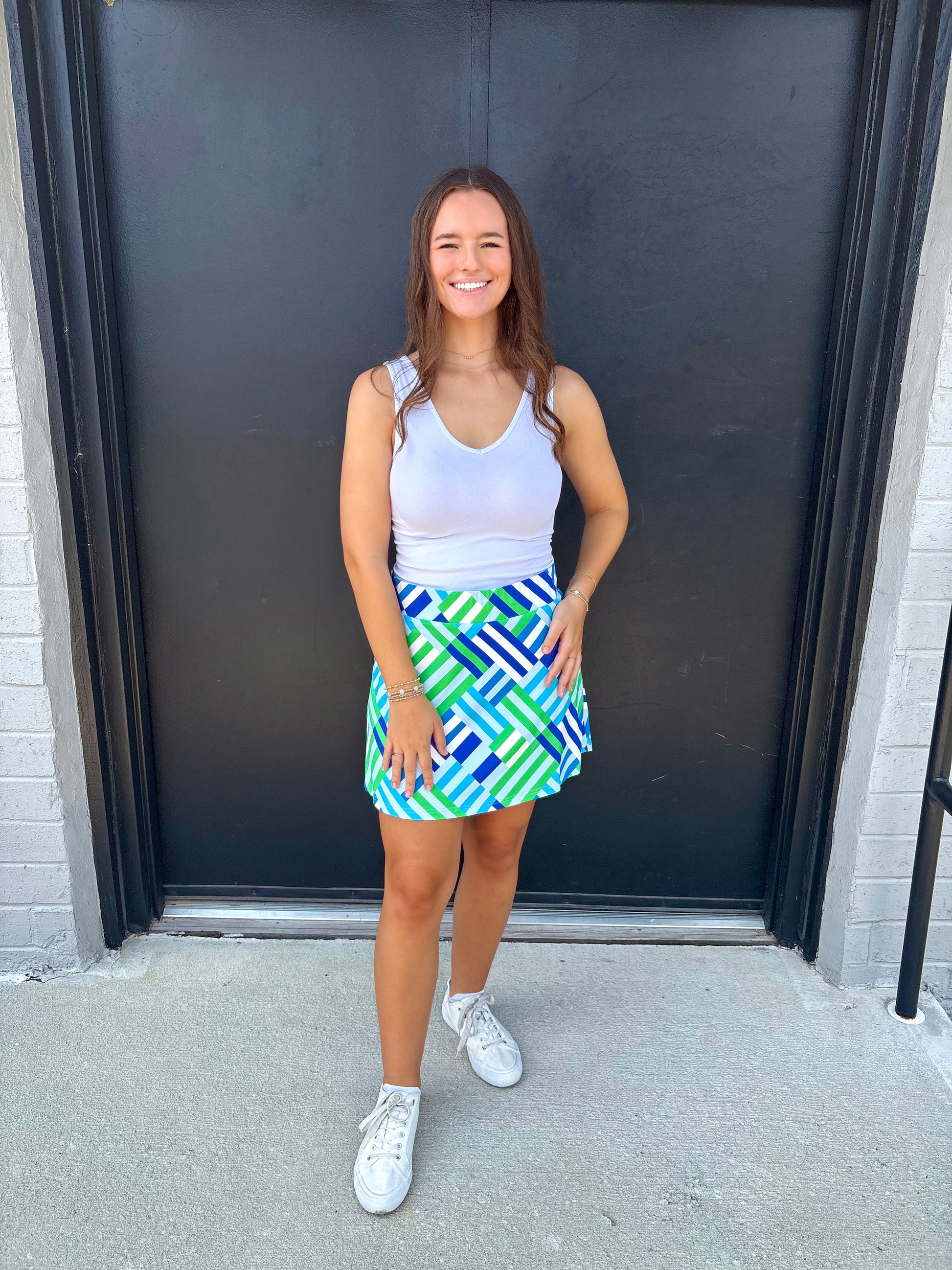 Woman wearing a white tank top and colorful geometric skirt standing in front of a black door.