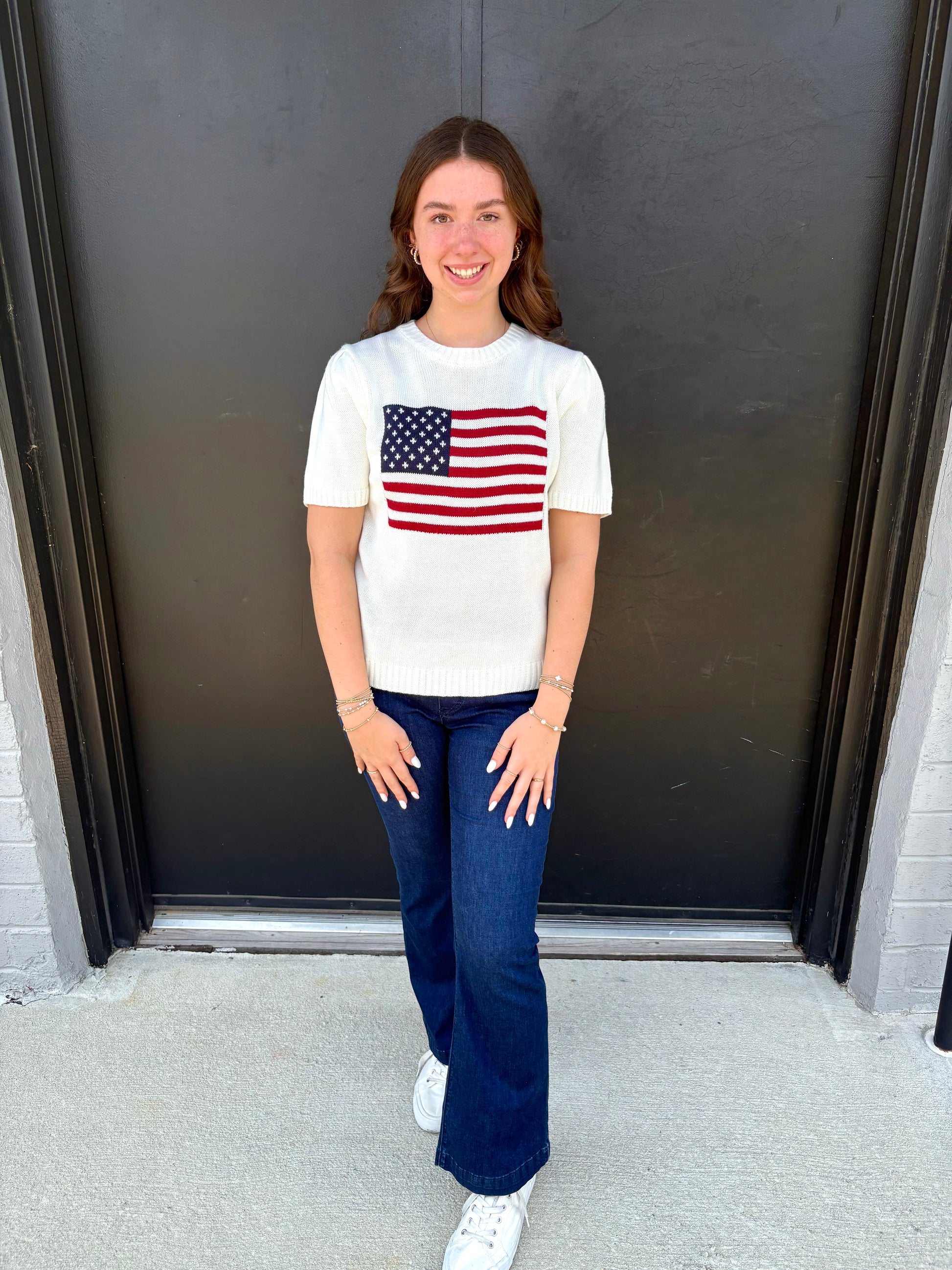 Person wearing a white sweater with an American flag design and blue jeans standing against a dark wall.