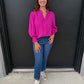 Woman wearing a bright pink blouse and blue jeans standing against a black wall.
