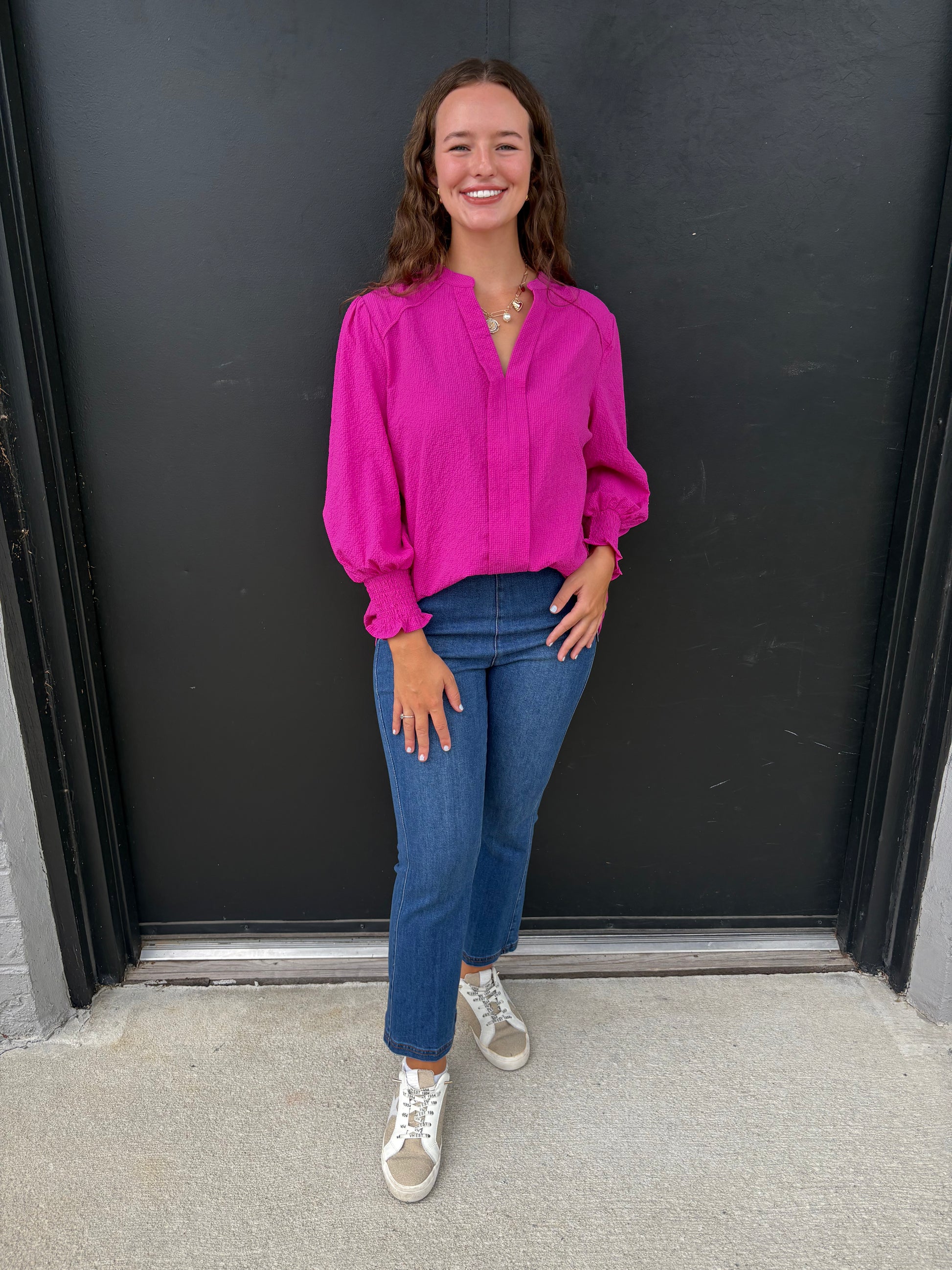 Woman wearing a bright pink blouse and blue jeans standing against a black wall.