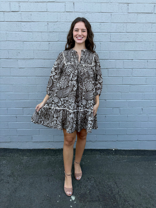 Woman wearing a patterned dress standing against a light blue brick wall.