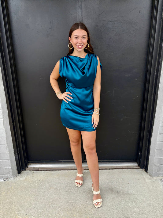 This is a sleek, jewel-toned teal satin mini dress with an elegant draped neckline and a figure-skimming silhouette.

Color: Rich teal/peacock blue, with a luminous satin finish that catches the light beautifully.
Neckline: Soft cowl neckline with dramatic draping across the chest for an elegant, fluid effect.
Sleeves: Sleeveless, with wide, softly gathered shoulder details that create a subtle cap-sleeve effect.
Bodice: Draped and relaxed through the bust, while the waist is more defined for a flattering s