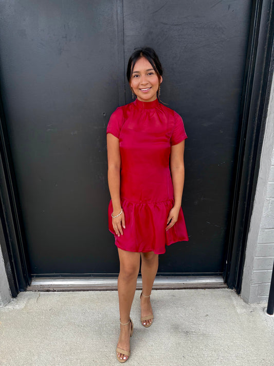 This is a sleek, feminine fuchsia mini dress with a sophisticated sheer neckline and softly flared skirt.

Color: Vibrant red/wine, giving the dress a bold, elegant presence.
Neckline: High mock neckline with a gathered, slightly ruffled finish.
Bodice: The upper chest features a sheer tonal mesh overlay, creating a subtle illusion neckline while maintaining a polished look.
Sleeves: Short, fitted sleeves with sheer fabric that coordinates with the neckline.
Waist: The bodice is fitted through