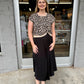 This is a chic leopard-print knit top with a feminine twisted-front detail and contrast trim.

Print: Classic leopard print in black, warm beige, and cream tones.
Neckline: Simple round crew neckline with a contrasting cream-colored ribbed edge.
Sleeves: Short, slightly relaxed cap sleeves, giving it an easy, polished shape.
Fit: Comfortable and gently fitted through the bodice without feeling overly tight.
Hem: The standout feature is the twisted/gathered center-front hem, creating a subtle sweetheart-like