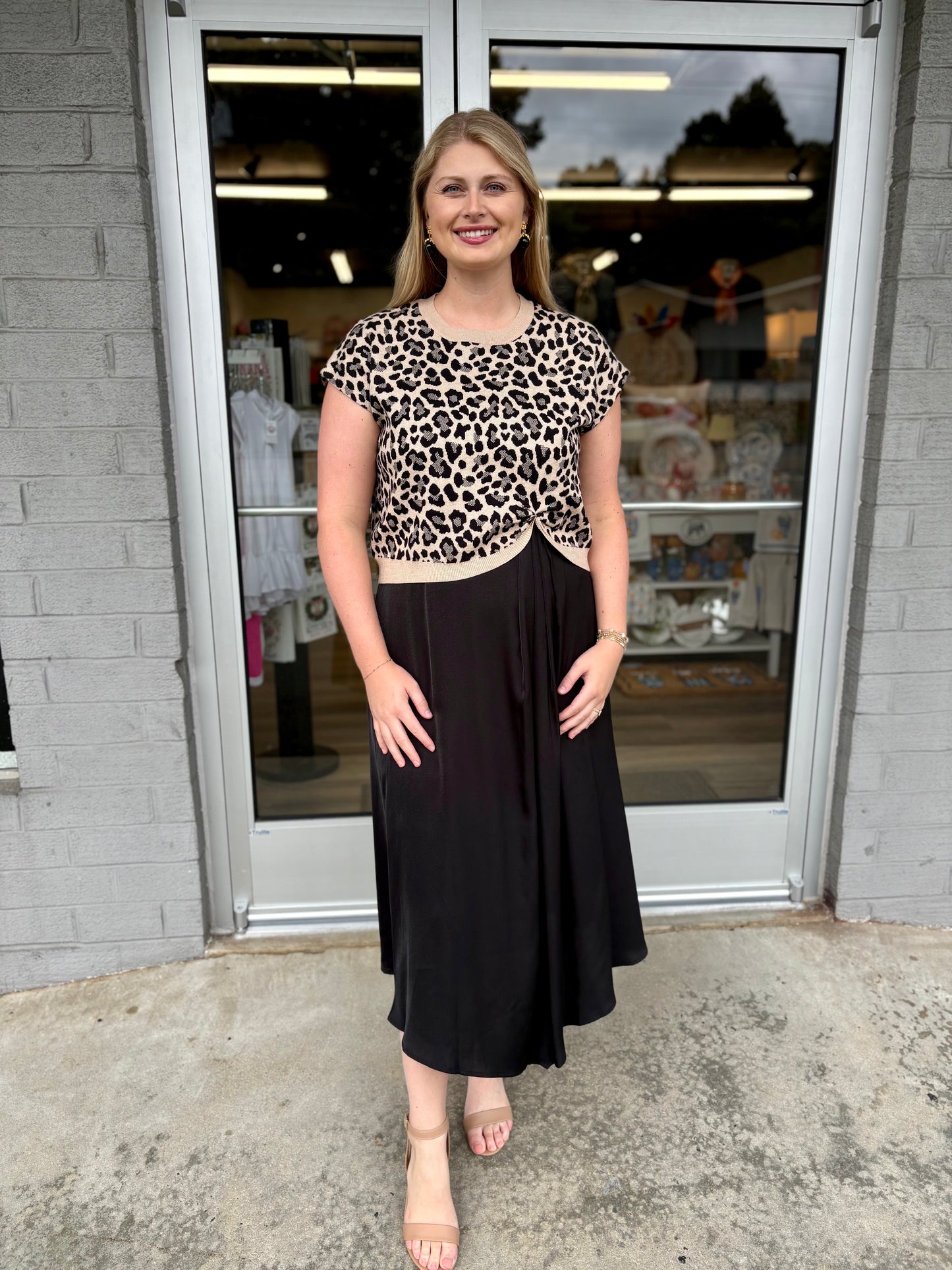 This is a chic leopard-print knit top with a feminine twisted-front detail and contrast trim.

Print: Classic leopard print in black, warm beige, and cream tones.
Neckline: Simple round crew neckline with a contrasting cream-colored ribbed edge.
Sleeves: Short, slightly relaxed cap sleeves, giving it an easy, polished shape.
Fit: Comfortable and gently fitted through the bodice without feeling overly tight.
Hem: The standout feature is the twisted/gathered center-front hem, creating a subtle sweetheart-like