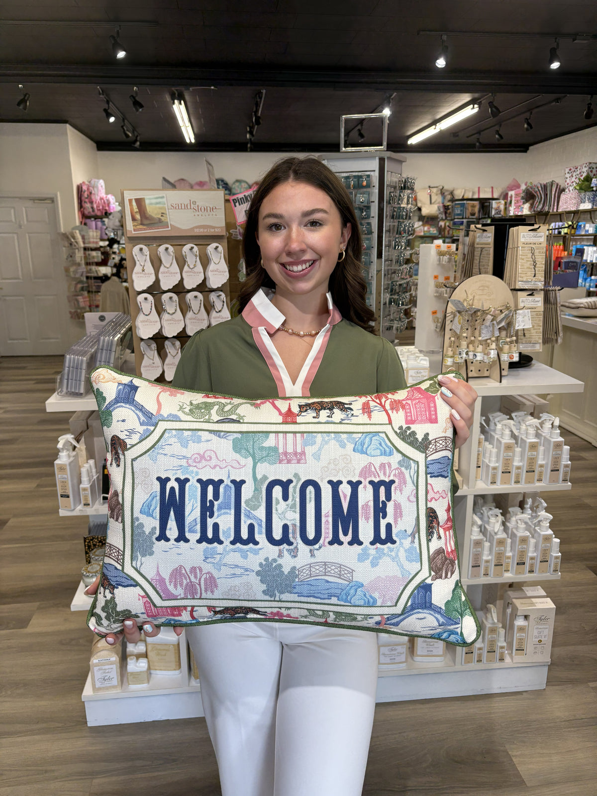 This decorative rectangular welcome pillow features a charming, colorful scenic print with whimsical illustrations of animals, trees, bridges, pagodas, florals, and landscape motifs in soft shades of blue, green, pink, red, and brown. The center showcases a large framed panel with “WELCOME” in bold navy lettering. Finished with green piping around the edges, this pillow has a playful yet polished look that adds a cheerful, inviting touch to an entryway, porch, living room, or guest space.