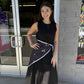 This chic black midi dress features a classic sleeveless silhouette with a round neckline and a fitted bodice that flows into a full, A-line skirt. The simple solid black design gives it a timeless, versatile look that can easily be dressed up or down. Styled here with a black-and-white polka dot scarf tied at the waist, it gets a fun, feminine pop of pattern and a flattering defined shape. Perfect for work, dinner, special occasions, or an elevated everyday look.