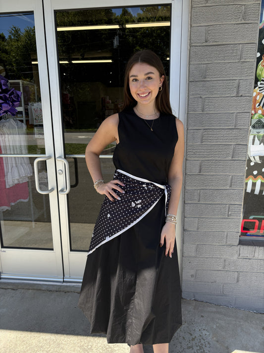 This chic black midi dress features a classic sleeveless silhouette with a round neckline and a fitted bodice that flows into a full, A-line skirt. The simple solid black design gives it a timeless, versatile look that can easily be dressed up or down. Styled here with a black-and-white polka dot scarf tied at the waist, it gets a fun, feminine pop of pattern and a flattering defined shape. Perfect for work, dinner, special occasions, or an elevated everyday look.