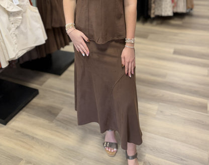 This is a chocolate brown maxi skirt with a soft, suede-like finish. It features a high-rise waist, fitted silhouette through the hips, and a gently flared A-line shape toward the hem. Subtle asymmetrical seam detailing across the front adds dimension, while the long, fluid cut gives it an elevated, minimalist look.