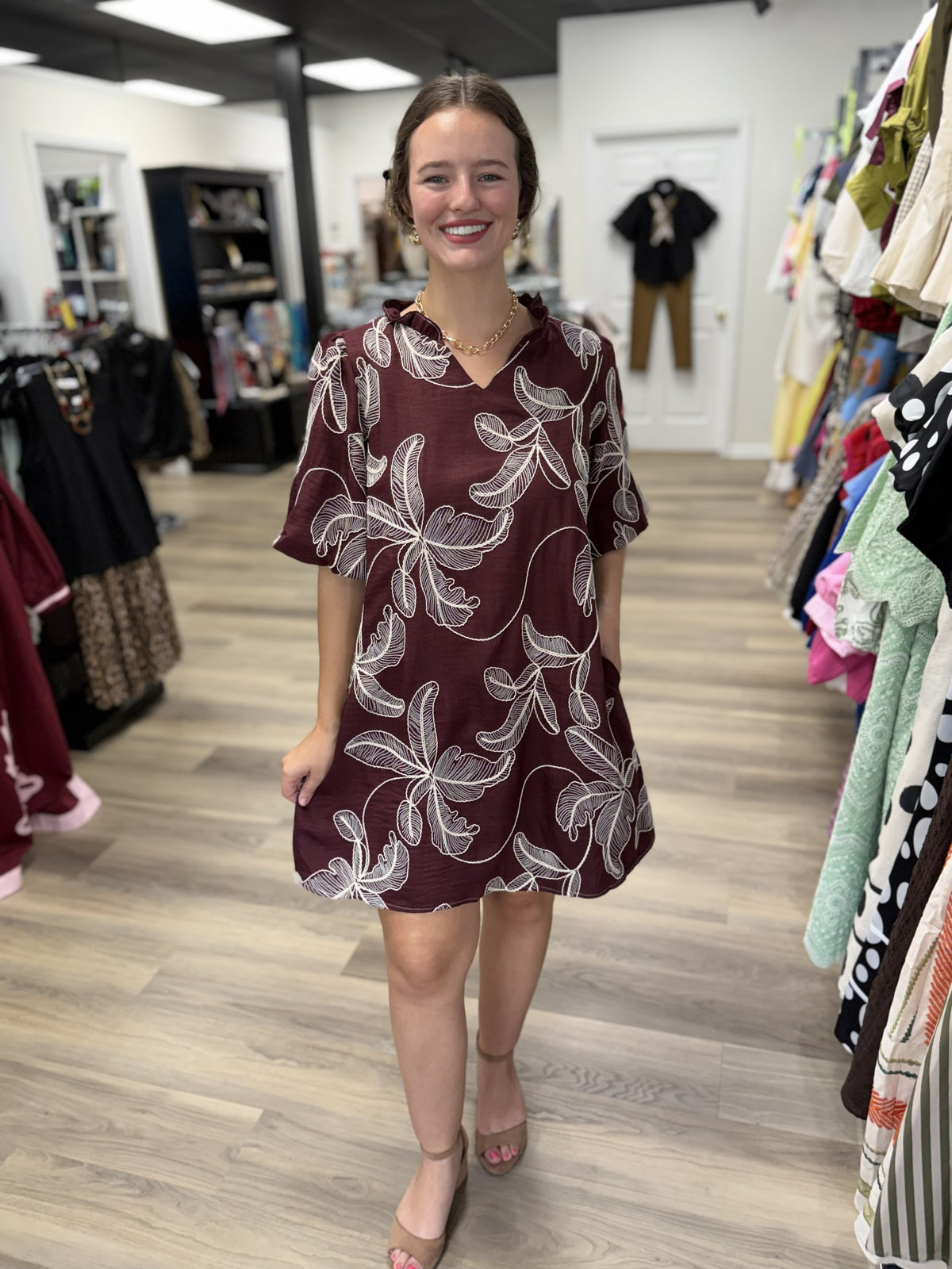 This dress has a rich, feminine boutique look with an easy, relaxed silhouette:

Deep burgundy/wine-colored base with an allover ivory botanical leaf print
V-neckline with a softly gathered, ruffled detail at the shoulders
Short, loose sleeves that create an airy, comfortable shape
Relaxed A-line silhouette that gently flares toward the hem
Above-the-knee length for a fresh, versatile look
Lightweight, textured-looking fabric that gives the dress a casual yet polished feel
The oversized botanical print adds