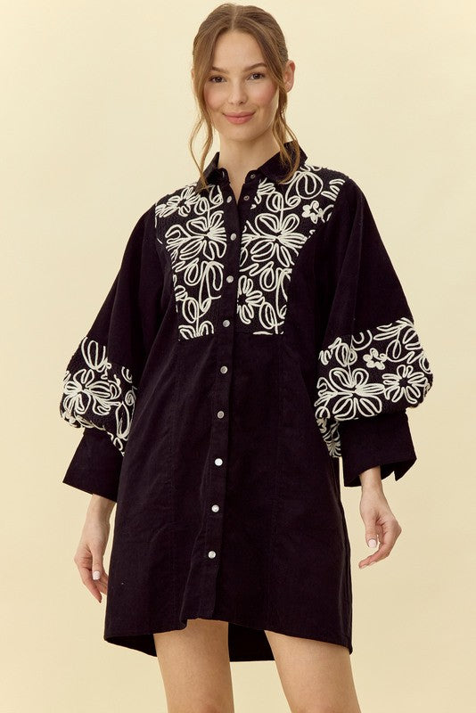 Short black button-up dress with white floral details around the chest and sleeves. Long balloon sleeves to a cuff