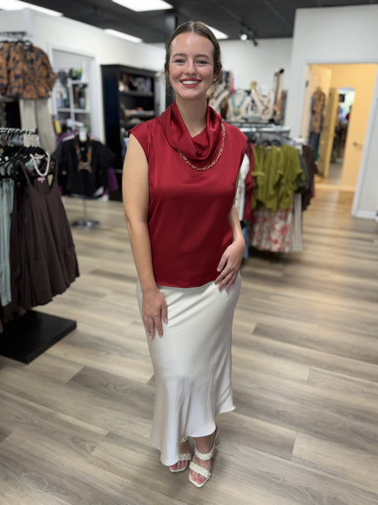 This top is an elegant deep burgundy satin blouse with a soft, lustrous finish. It features a sleeveless silhouette with extended shoulders and a beautifully draped cowl neckline that creates a sophisticated, fluid look. The relaxed, straight-cut bodice falls smoothly over the figure, while the rich wine-red color gives it a polished, dressy feel perfect for work, dinners, or special occasions.