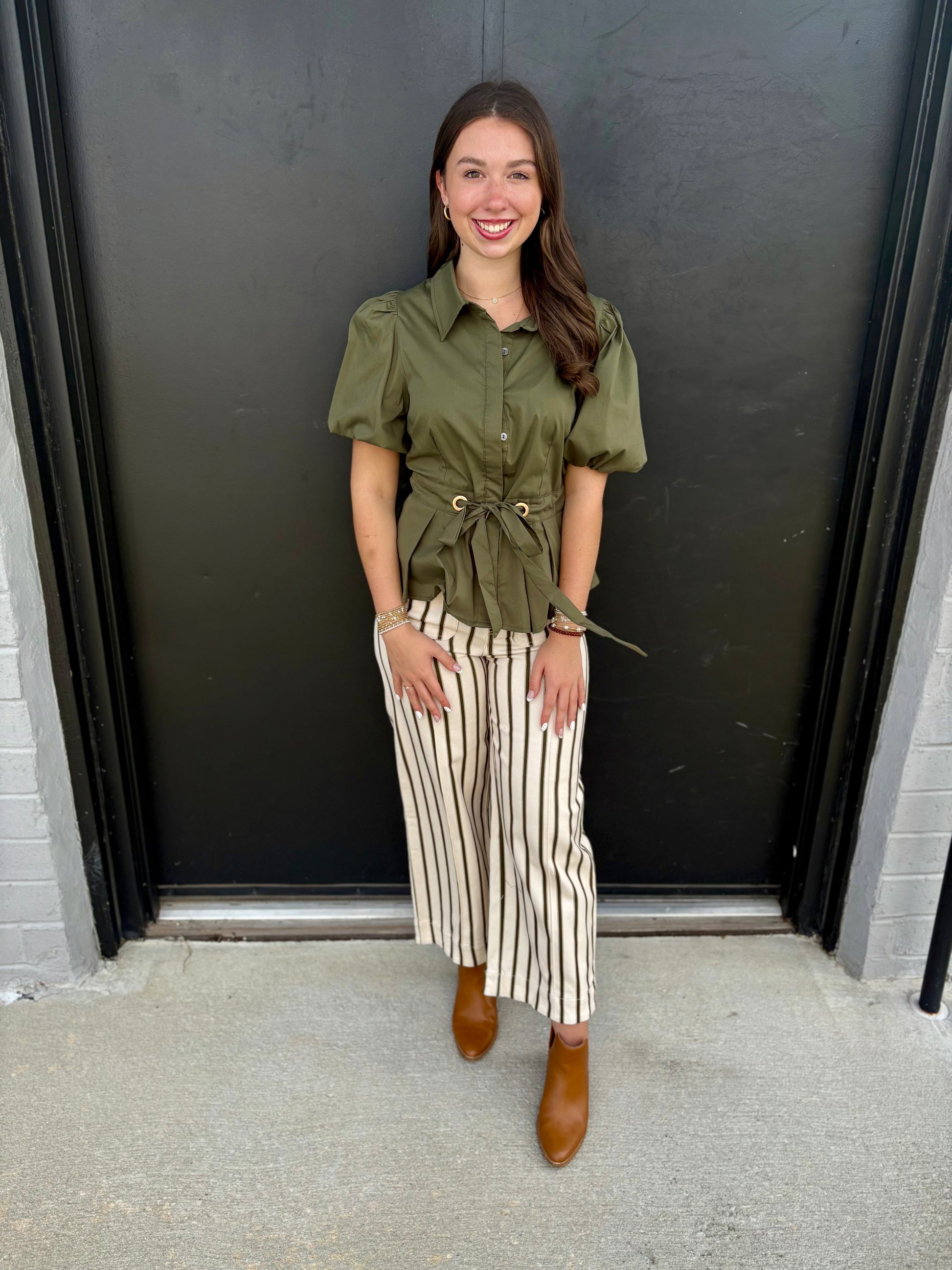 The pants are cream-colored cropped wide-leg trousers with a bold vertical stripe pattern.

Color: Warm ivory/cream base with thin dark brown or black vertical stripes.
Pattern: Classic pinstripe-style vertical stripes, creating a lengthening effect.
Fit: Relaxed through the hips and legs with a wide, straight-to-wide-leg silhouette.
Rise: Appears to be mid- to high-rise, sitting comfortably at the natural waist.
Length: Cropped, ending just above the ankle.
Fabric: Lightweight woven material with a softly 