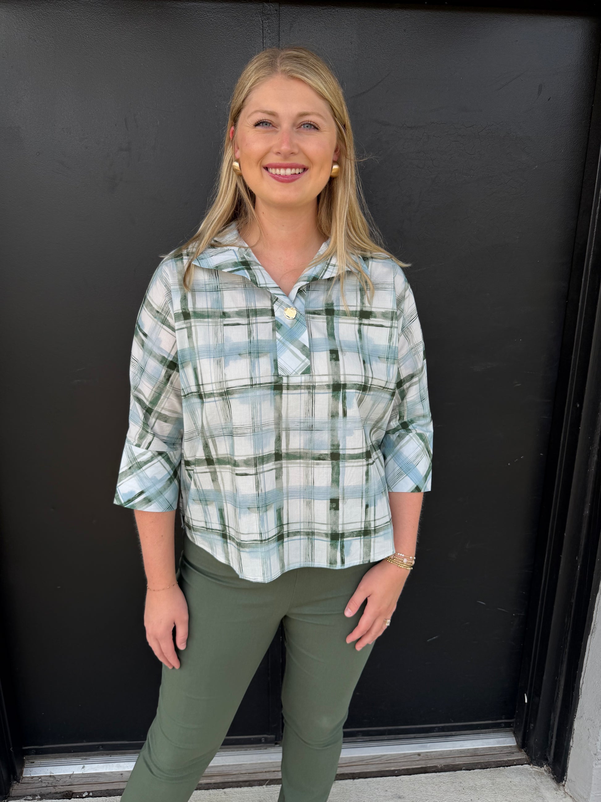 A relaxed, collared plaid pullover blouse with a polished casual feel.

Pattern: Soft plaid/check in sage green, muted blue, white, and gray.
Neckline: Wide, notched shirt-style collar with an open V-neck.
Front: Button-front styling, with a small gold-toned button visible near the chest.
Sleeves: Three-quarter length, slightly relaxed, with turned-back/cuffed-looking ends.
Fit: Loose and easy through the torso, with a gently curved hem that falls around the hips.