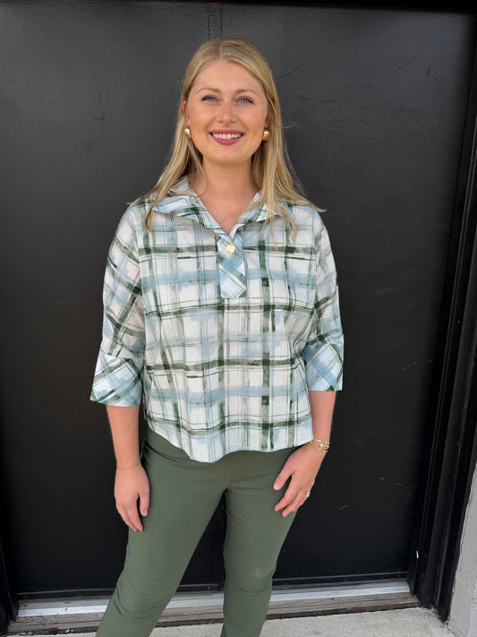 A relaxed, collared plaid pullover blouse with a polished casual feel.

Pattern: Soft plaid/check in sage green, muted blue, white, and gray.
Neckline: Wide, notched shirt-style collar with an open V-neck.
Front: Button-front styling, with a small gold-toned button visible near the chest.
Sleeves: Three-quarter length, slightly relaxed, with turned-back/cuffed-looking ends.
Fit: Loose and easy through the torso, with a gently curved hem that falls around the hips.