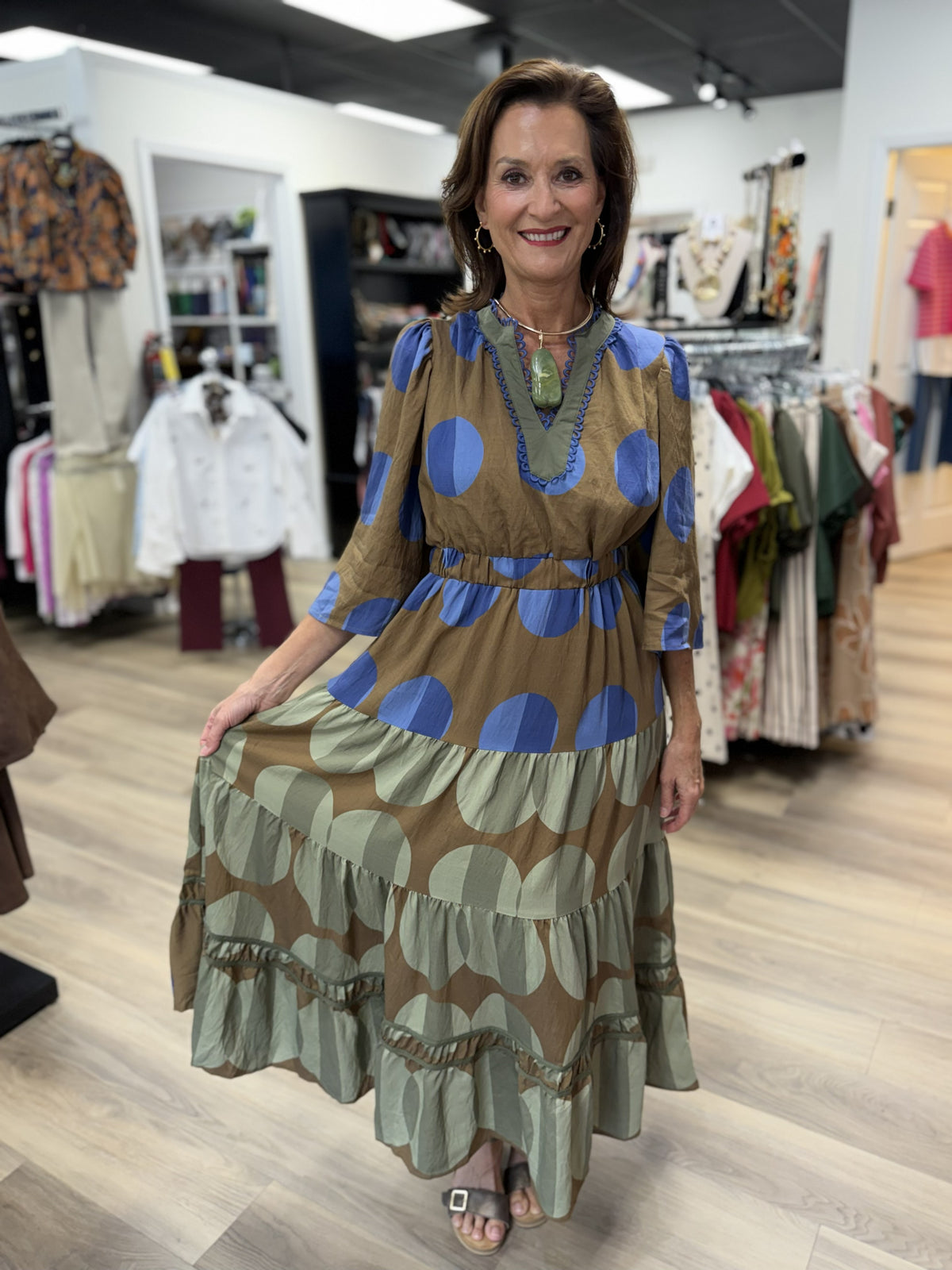 This is a boho-inspired maxi dress featuring an oversized polka-dot print in earthy brown, periwinkle blue, and soft sage green. It has a split neckline with contrasting sage trim and decorative blue scalloped detailing, along with gently puffed three-quarter sleeves.

A smocked elastic waist creates definition before flowing into a relaxed, tiered maxi skirt. The lower skirt transitions to a sage-and-brown oversized dot pattern and is finished with gathered tiers, decorative openwork trim, and a ruffled he