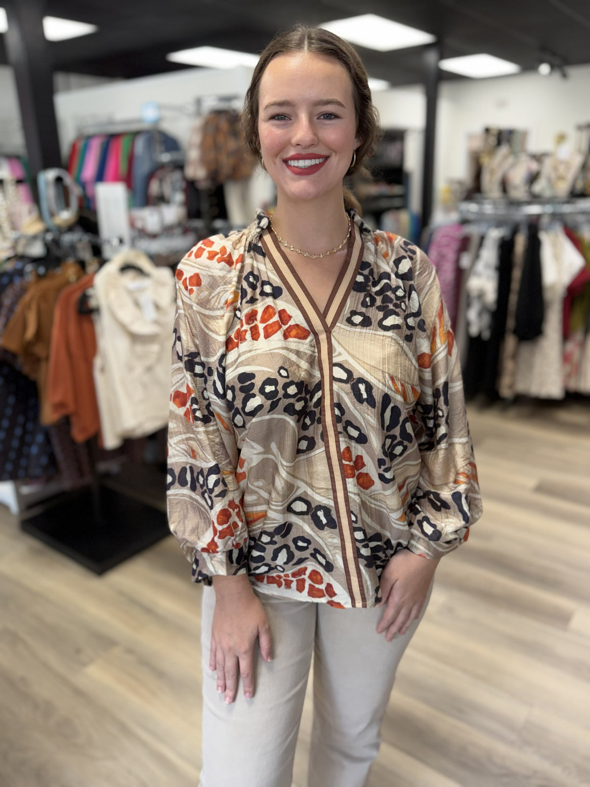 This is a flowy mixed animal-print blouse featuring an abstract combination of leopard-inspired spots and organic patterns in beige, taupe, cream, black, and burnt orange. It has a V-neckline with contrasting brown striped trim that continues vertically down the front for a polished accent. The blouse features a softly gathered neckline, relaxed fit, and voluminous three-quarter sleeves with gathered cuffs, giving it an easy, feminine silhouette.