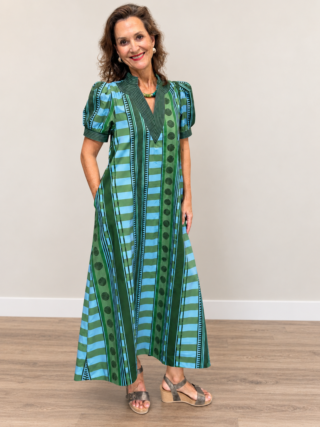 This is a green and sky-blue printed maxi dress featuring a bold mix of horizontal stripes, vertical geometric borders, and dotted motifs. It has a structured split V-neckline with contrasting striped trim and short puff sleeves finished with coordinating cuffs. The relaxed bodice flows into a full, A-line maxi skirt, creating an airy silhouette with plenty of movement. Side pockets add a practical touch, while the mixed-print design gives the dress a vibrant, artistic feel.