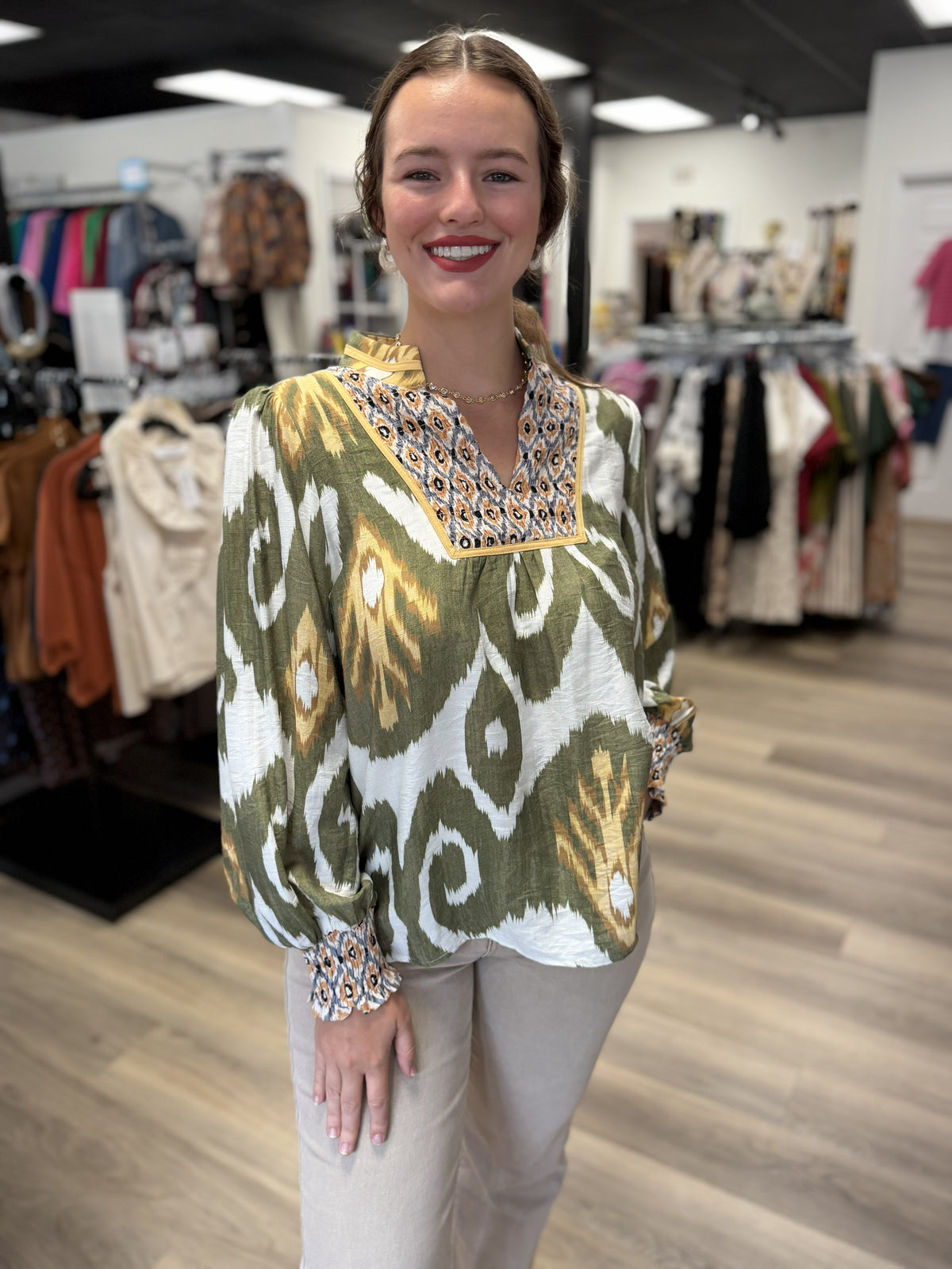 This top is a flowy olive green printed blouse featuring a bold ikat-inspired pattern in ivory, mustard, and golden tan. It has a split V-neckline with a short stand collar and a contrasting animal-print inset framed with mustard trim for an eye-catching mixed-print look. The long, softly gathered sleeves finish with smocked animal-print cuffs, while the relaxed silhouette and gently rounded hem give it an easy, comfortable drape.