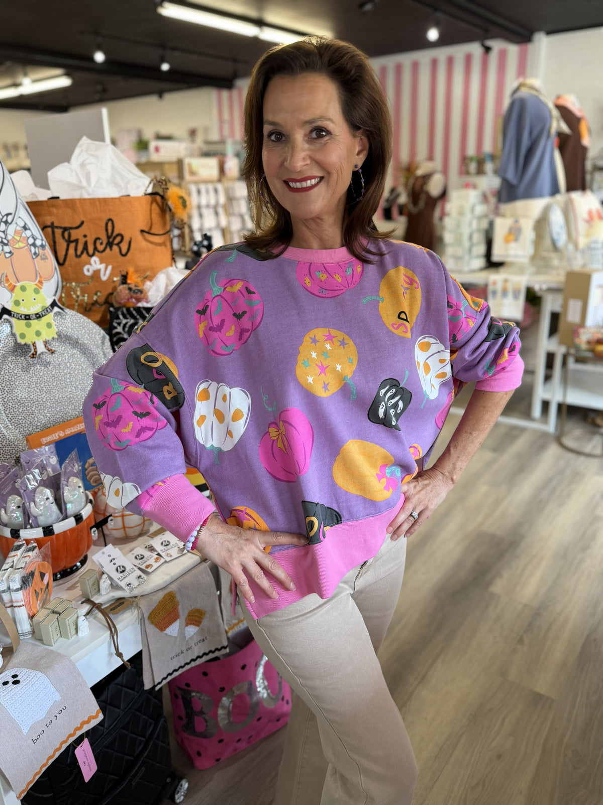 This is a purple Halloween-themed sweatshirt featuring a playful allover pumpkin print in bright pink, orange, white, and black. The pumpkins showcase a variety of whimsical patterns and spooky details, including bats, stars, ghosts, and colorful accents. It has a relaxed, oversized fit with dropped shoulders and long sleeves, finished with contrasting bright pink ribbed trim at the crew neckline, cuffs, and hem. The bold colors give this festive sweatshirt a fun, feminine twist on classic Halloween style.