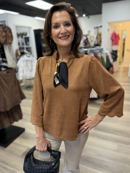 This rich camel brown top features a soft suede-like texture with an effortlessly polished silhouette. It has a feminine ruffled stand collar, subtle puff shoulders, and relaxed three-quarter sleeves finished with scalloped cuffs. A coordinating scalloped hem adds a charming touch, while the easy, slightly relaxed fit makes it comfortable and flattering. Styled with a printed scarf at the neckline, it’s a versatile fall favorite that pairs beautifully with denim or neutral pants.