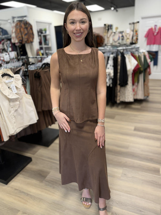 This outfit is a coordinating chocolate-brown two-piece set with a soft, suede-like finish. The sleeveless top features a square neckline, wide straps, vertical seam detailing, a fitted bodice, and a subtly flared hem. It’s paired with a matching high-waisted midi/maxi skirt that has a streamlined fit through the hips and gently flares toward the hem, with curved seam detailing adding shape and interest.